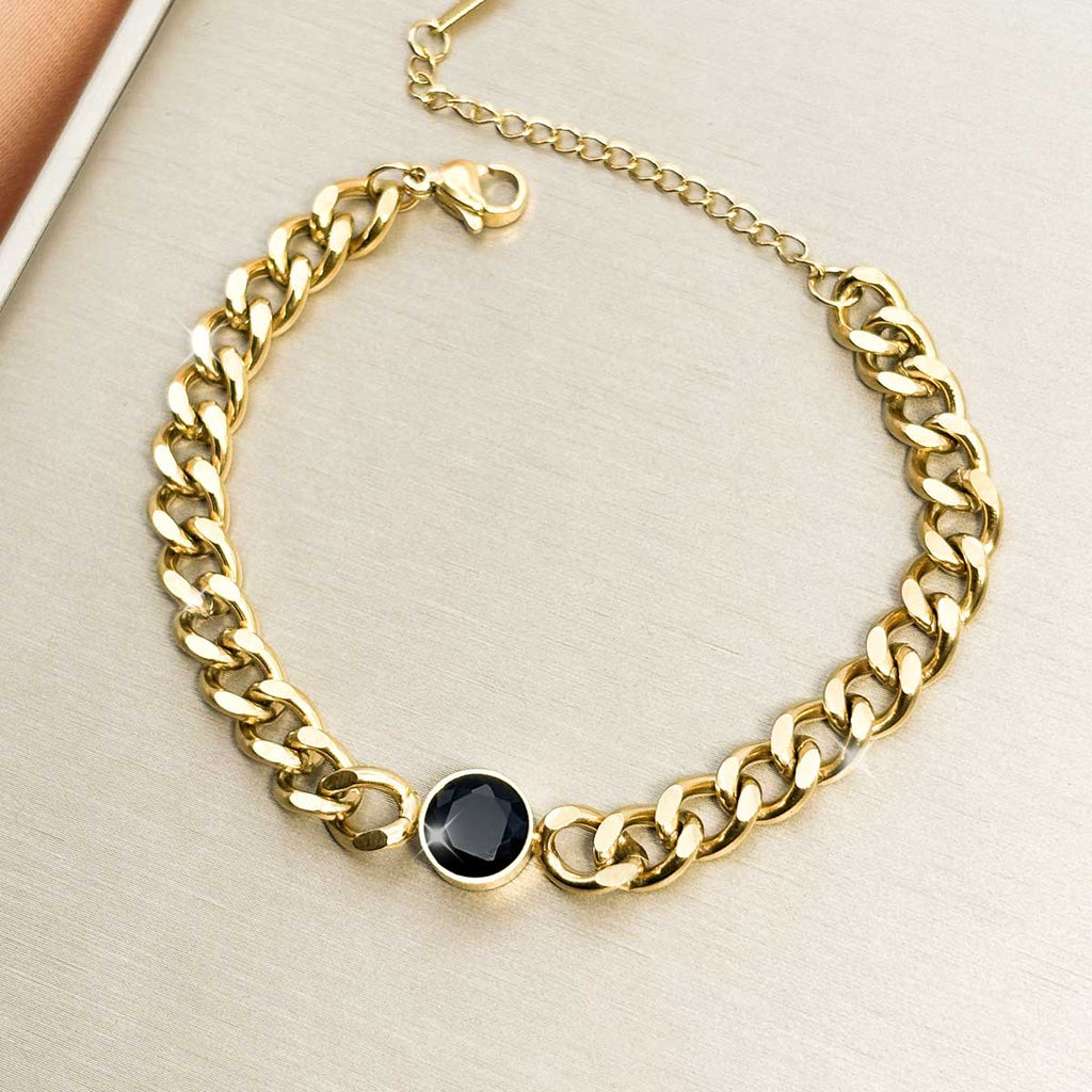 Goldfarbenes Edelstahl-Armband für Damen aus flachen Panzerkettengliedern und einem runden, schwarzen Element im Zentrum, Studioaufnahme.
