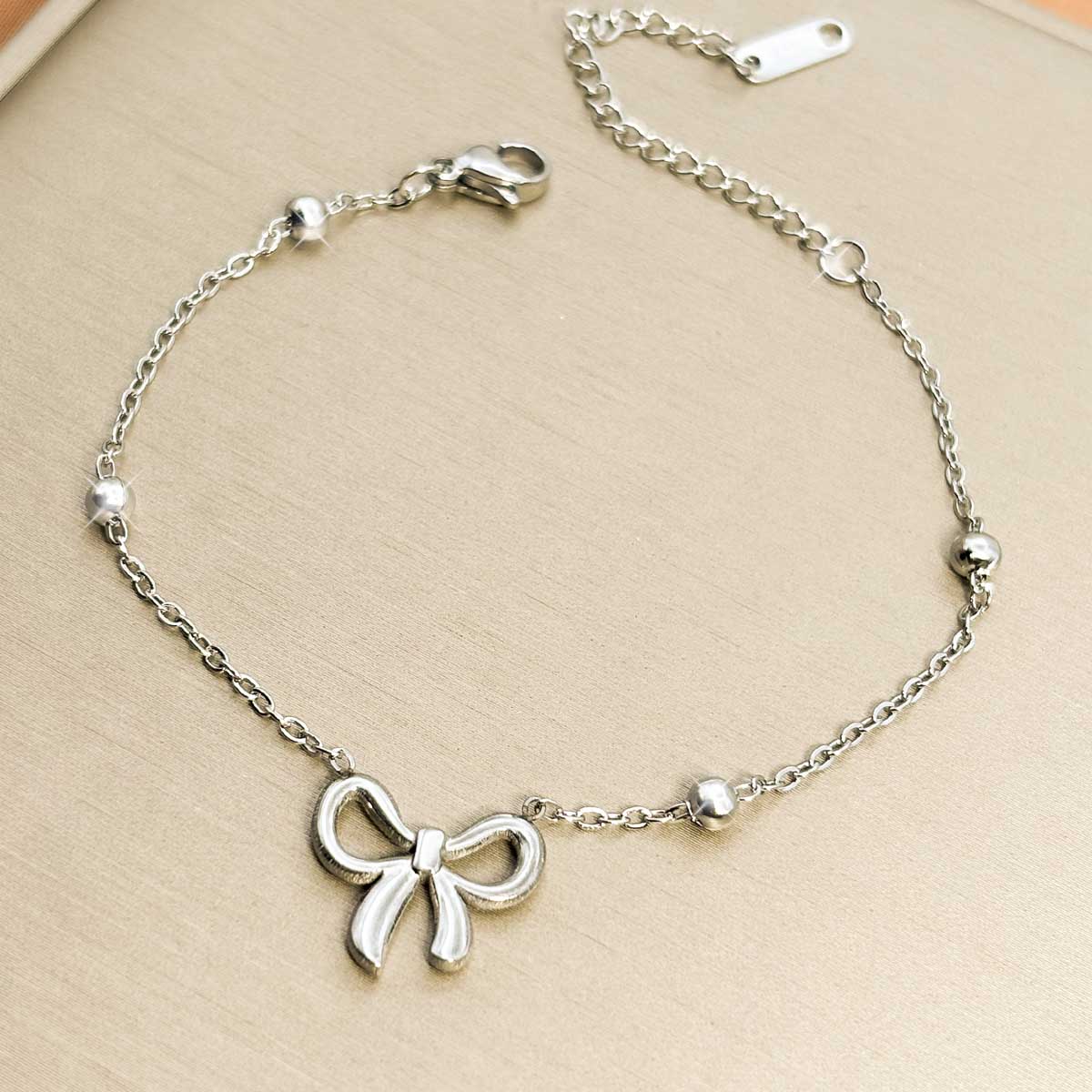 Silberfarbenes Edelstahl-Armband für Damen, dezent besetzt mit kleinen Kugeln und einem zarten Schleifen-Charm, Studioaufnahme.