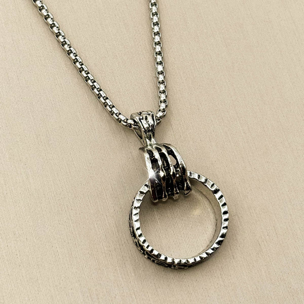 Silberfarbene Edelstahl-Herrenkette mit einem Anhänger in Form einer Hand, die einen strukturierten Ring greift, Studioaufnahme von links.