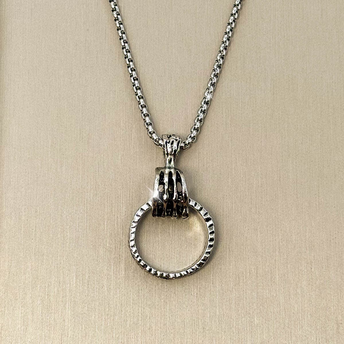 Silberfarbene Edelstahl-Herrenkette mit einem Anhänger in Form einer Hand, die einen strukturierten Ring greift, Detailaufnahme.